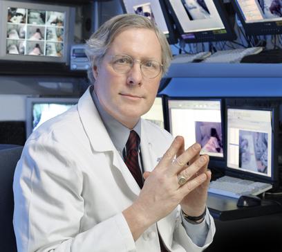 Dr. Gregory Bergey, in a white lab coat with his hands together in a lab with computer monitors behind him displaying various medical images.
